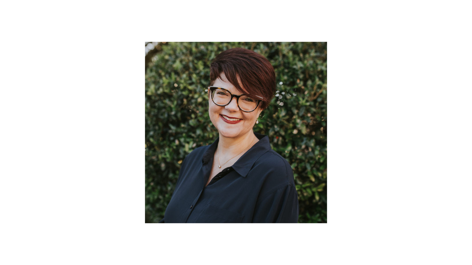 A woman with glasses standing in front of bushes.